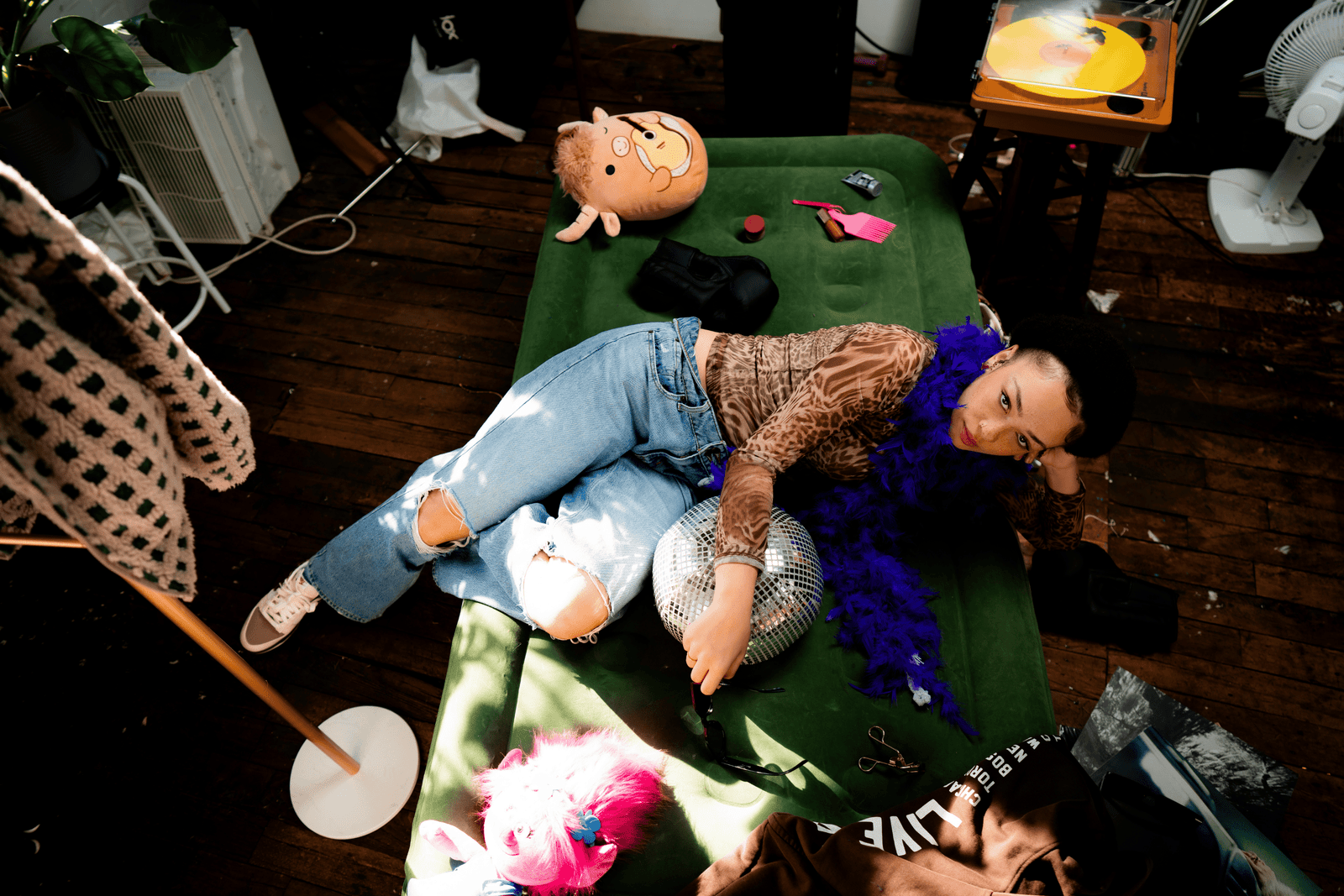 Woman in animal print shirt, blue feather boa and blue jeans on green inflatable mattress with miscellaneous items strewn around